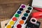 Angled view over a wooden desk with school supplies on it. Stepler, colored pencils, notebooks, calculator, tapes, paints. Back to