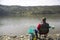 Angler With His Back Towards Camera Sitting On The River Coast Enjoying Angling And Beautiful Nature