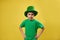 Angry boy wearing leprechaun green hat poses to the camera on a yellow background. Saint Patrick`s Day. Copy space