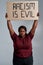 Angry plus size young african american woman in casual clothes looking at camera, holding Racism is evil banner above