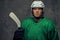 Angry professional hockey player in green sportswear standing with a hockey stick on a gray background.