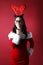 Angry young woman with reindeer attire and glasses and santan in santa helper hat with gift box front of red background