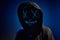 Anonymous man in hood hiding face behind neon glow scary mask on dark background. Horror concept