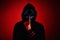Anonymous man in hoodie hiding face behind neon glow scary mask on red background. Horror concept