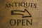 Antiques Open sign set on a wood background and a right facing arrow. Brown 3D text against wood grain background.