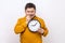 Anxious nervous middle aged man biting nails on his fingers holding big wall clock in hand.