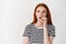 Anxious redhead girl biting finger and looking doubtful left, frowning worried, standing over white background