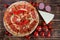 Appetizing pizza with salami and chilli pepper lies on a wooden surface next to a special circular knife, a piece of cheese,