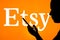 April 22, 2022, Brazil. In this photo illustration the Etsy logo seen in the background of a silhouetted woman holding a mobile