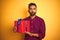 Arab indian hispanic man holding birthday gift standing over isolated yellow background with a confident expression on smart face