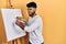 Arab man with beard standing drawing with palette by painter easel stand angry and mad screaming frustrated and furious, shouting