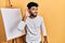 Arab man with beard standing by painter easel stand smiling with hand over ear listening and hearing to rumor or gossip