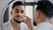 Arabian indian arab hispanic millennial 30s bearded man applying moisturizer cream on face skin looking in bathroom