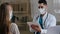 Arabian male doctor physician in medical face mask making notes on clipboard consulting female patient explaining