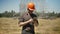 Architect in helmet and glasses using tablet and standing on straw field, metal construction on background, concentrated