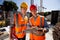 Architect and structural engineer dressed in orange work vests and helmets discuss a building project on the tablet on