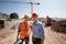 Architect and structural engineer dressed in orange work vests and helmets stand on the open air building site