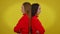 Argued young twin sisters standing back to back at yellow background with displeased facial expression. Side view