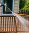 Arm holding a water pressure sprayer is seen spraying water on the brown wooden railing of a deck.
