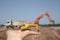 On the arm is a small orange toy excavator. Behind him, out of focus, is a real working excavator and dump truck