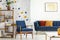 Armchair and couch with pillows in blue and orange living room interior with poster and plant. Real photo