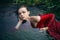 Art beautiful romantic portrait of a sexy young woman in a red dress lying in a river with green algae in summer in