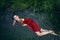 Art beautiful romantic portrait of a sexy young woman in a red dress lying in a river with green algae in summer in