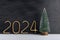 Artificial small Christmas tree on wooden stand and golden number 2024 on black background. New year background