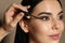 Artist applying black eyeliner onto woman`s face on beige background, closeup