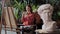 Artist in an art studio with plants - young pretty woman takes a photo of her painting - greek head sculpture on a