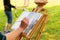 Artist painting on open air, wooden easel sketchbook with unfinished painting close-up. Female hand paints in a sunny summer