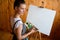 Artist stands near an easel with palette of multi-colored paints in her hands.