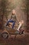 Artistic outdoor portrait of two blond girls playing with a wheel barrow in a woods