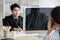 Asian beautiful female employee in black and white stripe shirt sit on chair smiling talk on phone while male worker on opposite