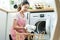 Asian beautiful woman put dirty clothes to washing machine in house. Attractive girl wear apron sit on floor, feel happy to loads