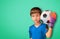 Asian boy is football goalkeeper wearing gloves and holding a soccer ball on green background copy space