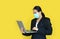 Asian business woman wearing medical shielding mask working with laptop computer isolated on yellow background