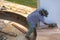 Asian carpenter using electric planer to shaving curved wooden bench surface at outdoor workshop