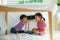 Asian cute little boy feeding snack to his sister sibling while sitting in a blanket fort in living room at home for perfect