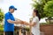 Asian delivery young man in blue uniform smile and holding a cardboard boxes in front house and Asian woman accepting a delivery