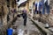 Asian elderly villager pours water from bucket on narrow street.