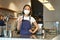 Asian female barista, girl in medical mask, shows okay sign, works in cafe behind counter, brews coffee, works with