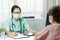 Asian female Doctor in green uniform wear eyeglasses and surgical mask writing something in checklist document while talking with