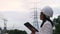 Asian female electrical engineer working on laptop near high voltage pole, inspecting power grid.