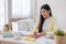 Asian female freelancer reading report on laptop and jot down note on table in bedroom at home.Work at home concept.work from home