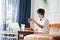 Asian female freelancer sitting on couch taking photo with smartphone & working on laptop at home