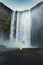 Asian female tourist in yellow jacket enjoying the Skogafoss waterfall flowing from cliff on summer in Iceland. Icelandic famous