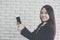 Asian girl smiles and uses her hand to point smartphone, background white brick wall, Online Trading Concepts and Auctions