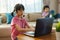 Asian girl student video conference e-learning with teacher and classmates on computer and her sibling reading book in sofa in