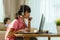 Asian girl student video conference e-learning with teacher and classmates on computer and her sibling reading book in sofa in